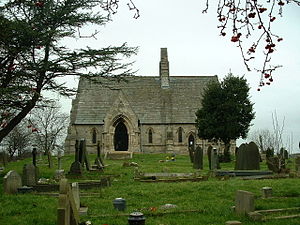 A small stone church seen from the south, with an extensive roof, small lancet windows, a prominent gabled porch, and a central bellcote
