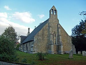 The west end of a stone church seen from a slight angle, showing two large buttresses with a window between, a double bellcote above, and part of the body of the church with its thatched roof