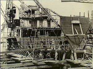 The stern of a ship undergoing repairs out of the water. The interior of the ship is exposed and cranes and workmen are working on the ship.