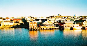  A cluster of buildings and wharves, with a small ship moored to the right of picture. The waterside buildings are clearly reflected in the sea.