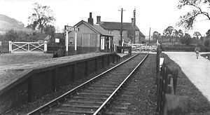 Small wooden railway station with a single rail track. The platform is considerably taller at one end than at the other. Aside from a small wooden building on the platform, the only other visible building is a single farmhouse.