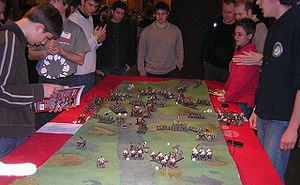 Several men surround a table, on which lies a small scale model of an undulating ground.  Many small figures of monsters and medieval warriors and siege weapons are placed on that terrain.  Dice are scattered among the figures.
