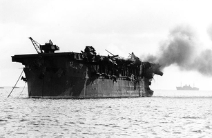Photograph of badly damaged aircraft carrier, afloat with smoke coming from deck.