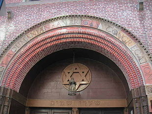 The top of an arched reddish-brick entrance-way is visible. Carved into stones on the top row of the arch are the words "First Roumanian-American Congregation", all in capital letters. The arch surmounts a brown wall with a bronze Star of David on it, with a lamp hanging from the arch in front of it. Underneath the brown wall, and above the doors, are inscribed the words "Shaarey Shamoyim" in Hebrew.
