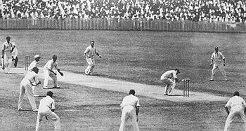 On a cricket field, a batsman ducks under the ball, watched by the man who had bowled the ball, another batsman and an umpire. There are a ring of men standing fielding next to the batsman. Everyone is wearing white shirts and trousers, with the fielders in caps. A large crowd is behind a fence in the background.