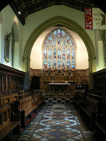 The same chapel with a wider chancel arch, decorated tiles, and a stone reredos below a large stained glass window. A flag with a ship on it hangs from high on the right wall