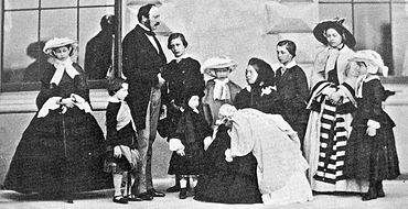 Photograph of a seated Victoria, dressed in black, holding an infant with her children and Prince Albert standing around her.