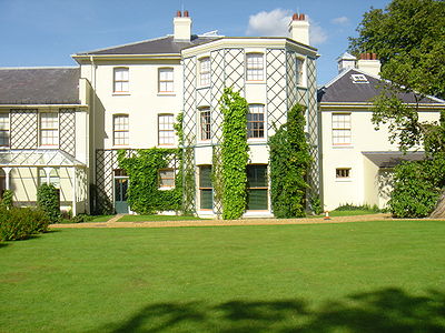 Stucco rendered building in a plain Georgian style with hipped slate roofs and overhanging eaves. The central block is three storeys, and these are two storey extensions on each side. Lattices are fixed to walls between windows to support climbing plants. A wide lawn forms the foreground, and tall trees appear behind the block to the right.