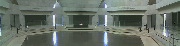 Panoramic view of the Hall of Remembrance. Hexagonal room with red-tile floor, limestone walls, and black panels. Eternal flame in foreground supported by a black box containing ashes from European Concentration Camps.