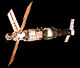 An image showing a white insulation covered, stepped-cylindrical module with a number of docking ports visible on a sphere at one end and three solar arrays projecting from the narrower portion of the stepped cylinder. A second, shorter module covered in darker insulation is docked to the end opposite that of the sphere, and a Soyuz spacecraft is docked to the other end of this module. The blackness of space serves as the image backdrop.